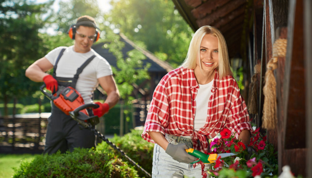 Strong caucasian man in uniform using electric trimmer for pruning bushes at garden while charming woman cutting dry leaves on flower in pots. Seasonal work outdoors.