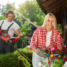 Strong caucasian man in uniform using electric trimmer for pruning bushes at garden while charming woman cutting dry leaves on flower in pots. Seasonal work outdoors.